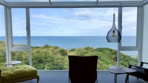 A room with a large window overlooking the ocean, a yellow armchair, a dark chair, and a hanging light fixture. - Twilight BnB (New Taipei City)