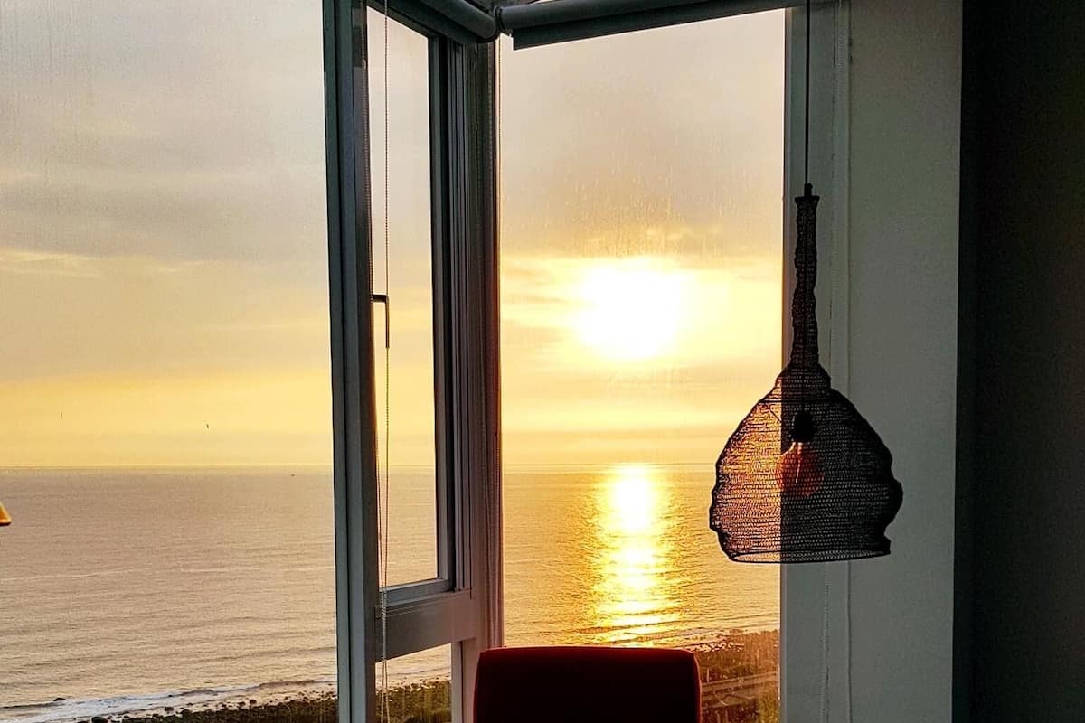 Une pièce dotée d’une grande fenêtre donnant sur un coucher de soleil sur la mer, d’une chaise et d’une lampe suspendue.