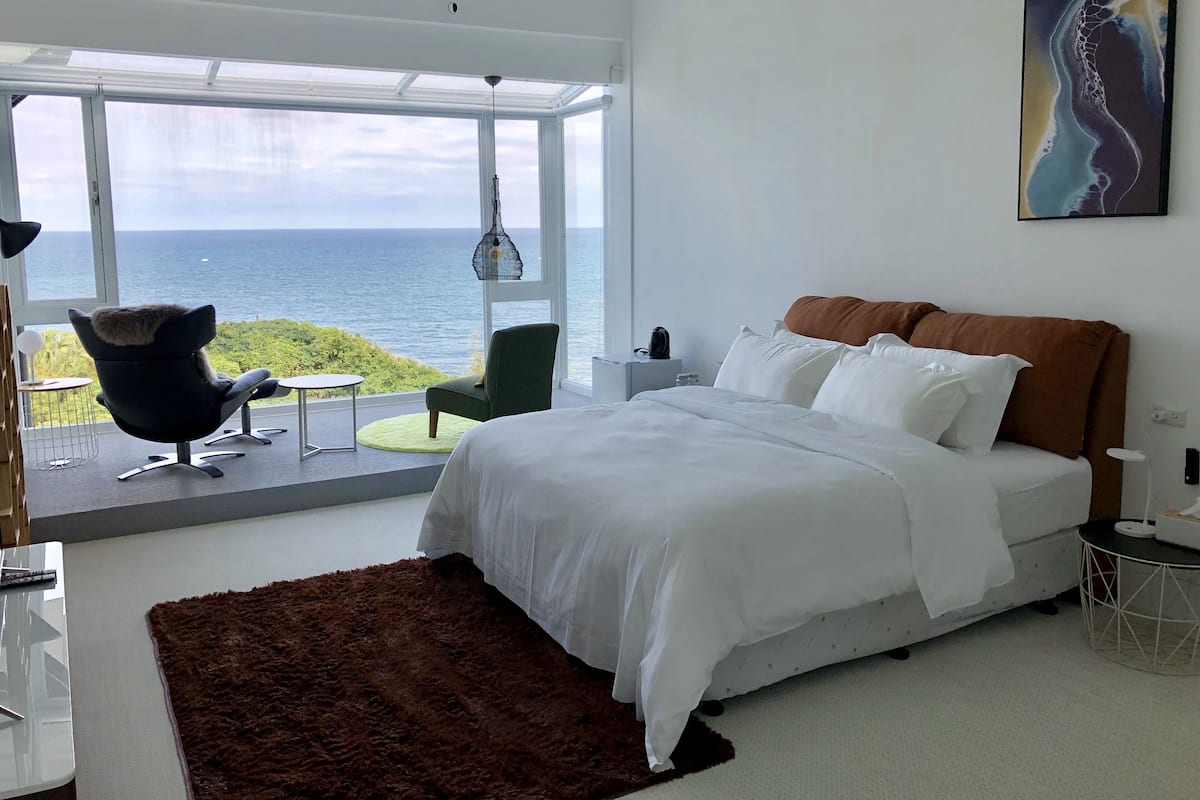 Une chambre avec un grand lit, une vue sur la mer, un fauteuil vert et un fauteuil noir près d’une fenêtre.