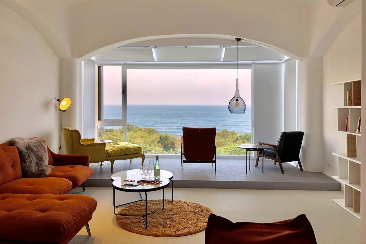 Un salon moderne doté d’une grande fenêtre donnant sur la mer, comprenant un canapé, des fauteuils, une table ronde et un luminaire suspendu.