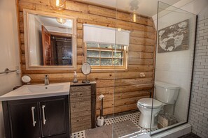 A bathroom with wooden walls, a white sink, a mirror, a toilet, and a glass shower enclosure. - Little Bear Cabin by Hauserman Rentals (Tahoe Vista)