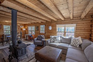 A log cabin interior with a stone fireplace, wooden ceiling beams, a gray sofa, and a stone stove. - Little Bear Cabin by Hauserman Rentals (Tahoe Vista)