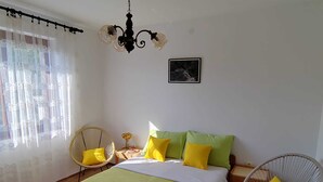 A bedroom with a bed, two chairs, a nightstand, a chandelier, a window with sheer curtains, and a framed picture on the wall.