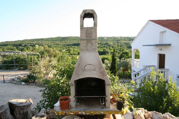 An outdoor stone oven with a chimney, surrounded by greenery and a white building with a red roof. - Apartment Vrban lijevi (Silo - island Krk)