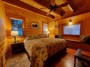 A cozy wooden cabin bedroom with a bed, two bedside lamps, a ceiling fan, and a window with blinds.