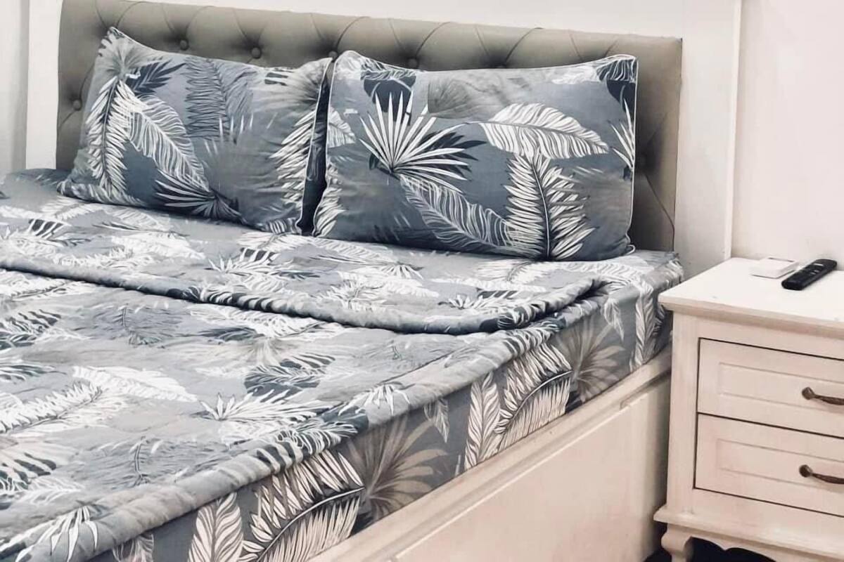 A bedroom with a bed featuring grey and white leaf-patterned bedding, a white headboard, and a bedside table with a remote control.