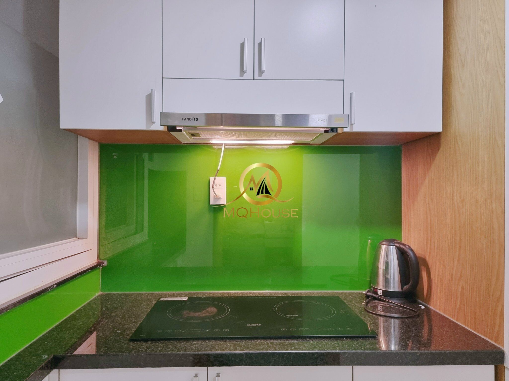 Een keuken met witte kastjes, een groene spatwand en het logo van MQHOUSE.