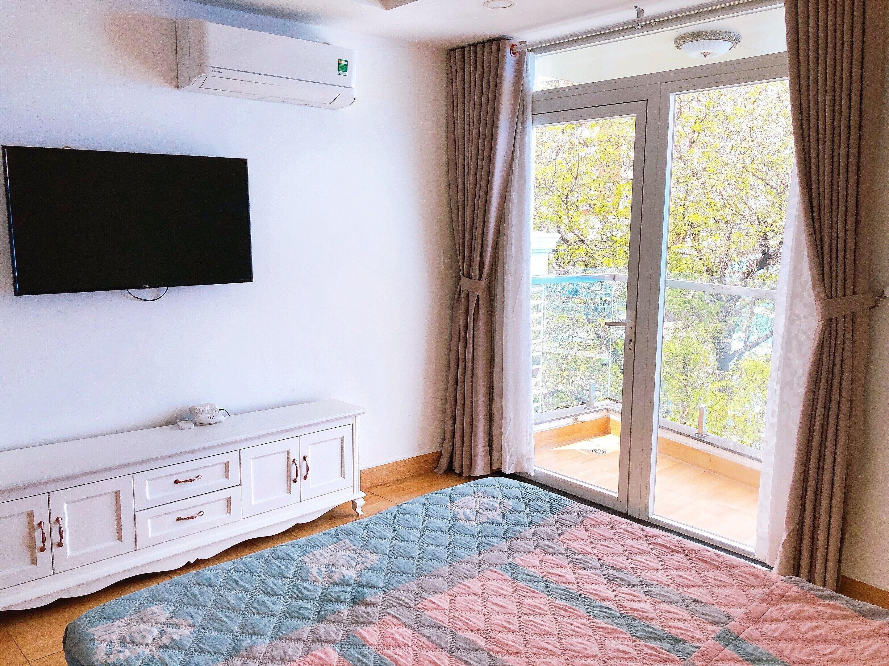 A bedroom with a large window, a flat-screen TV, a white dresser, and a colorful quilt.