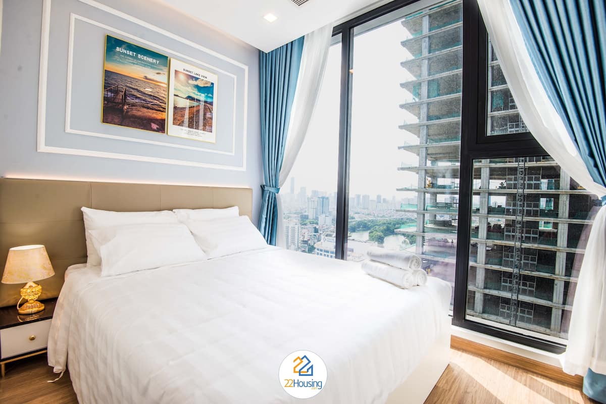 Ein modernes Hotelzimmer mit einem großen Bett, einem Nachttisch mit Lampe, Blick auf die Stadt durch ein großes Fenster und gerahmten Kunstwerken an der Wand.