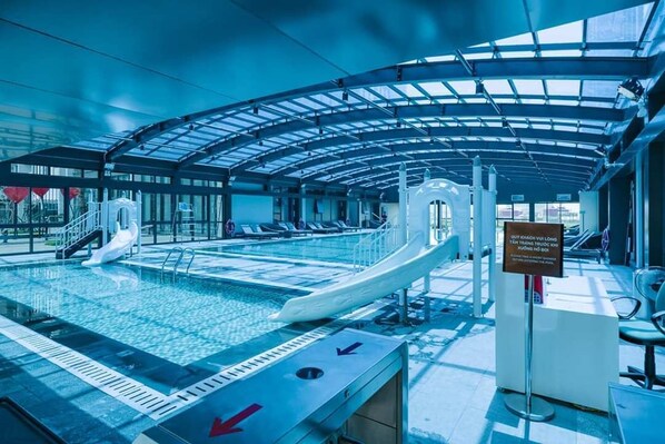 A modern indoor pool with a glass ceiling, a water slide, and clear signage.