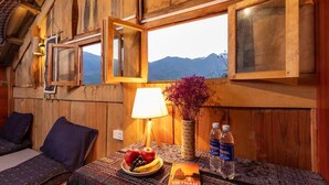 A wooden room with a bed, a table with a lamp, a book, and a view of mountains through a window. - Hmong Sister House - Hostel (Ta Van)