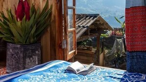 A room with a bed, a wooden wardrobe, a window with a view of a mountain, and a red patterned curtain. - Hmong Sister House - Hostel (Ta Van)
