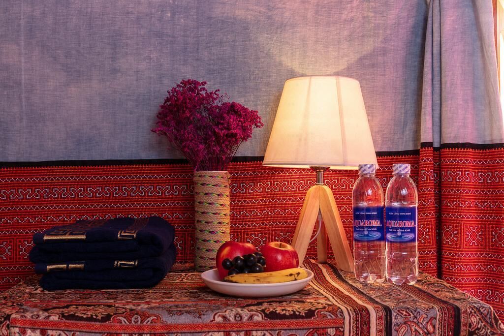 A hotel room with a patterned red carpet, a lamp, a plate of fruit, two bottles of Aquarona, a folded towel, and a vase with flowers.