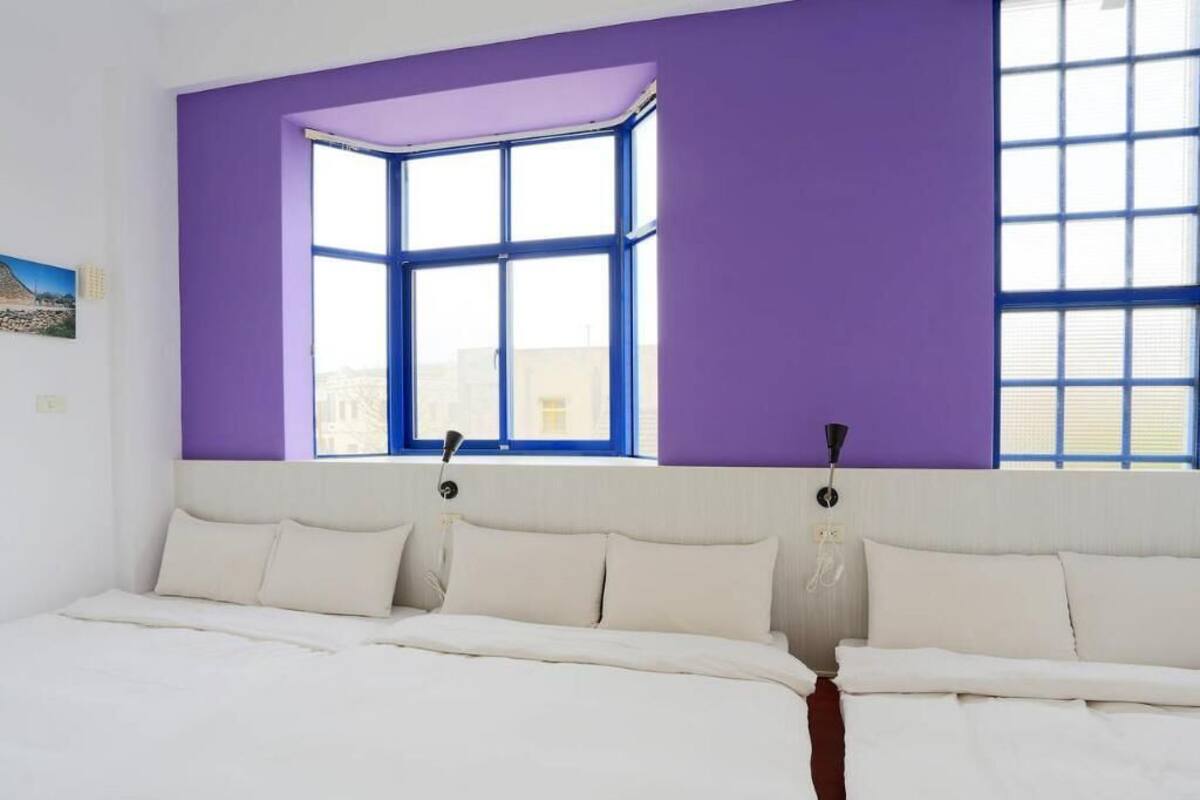 A bedroom with a large window, a bed with white linens, and a purple accent wall.