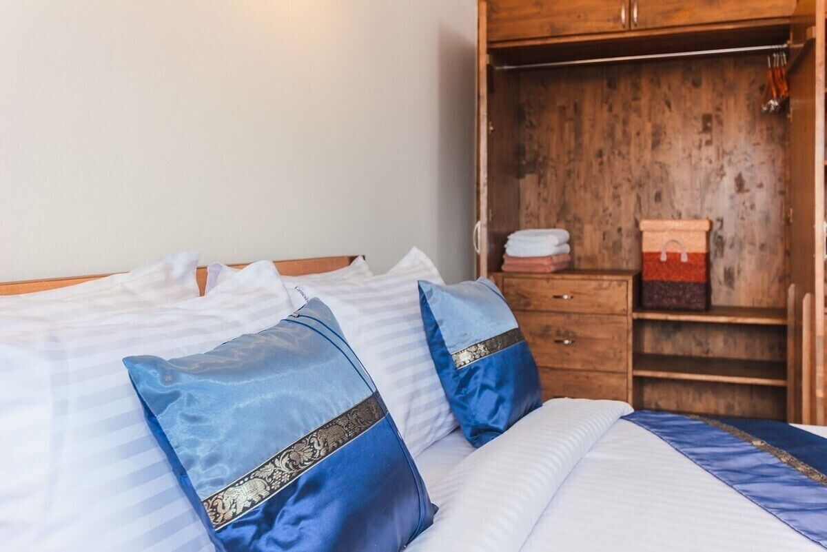 Una cama con ropa de cama blanca y azul, un armario de madera con espejo, y una estantería con toallas dobladas.