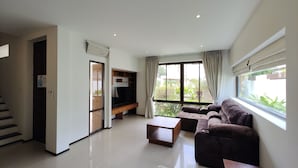 Una sala de estar moderna con un sofá, una mesita de centro y un mueble para el televisor. Se ve una escalera que sube a otro nivel, y ventanas amplias con cortinas.