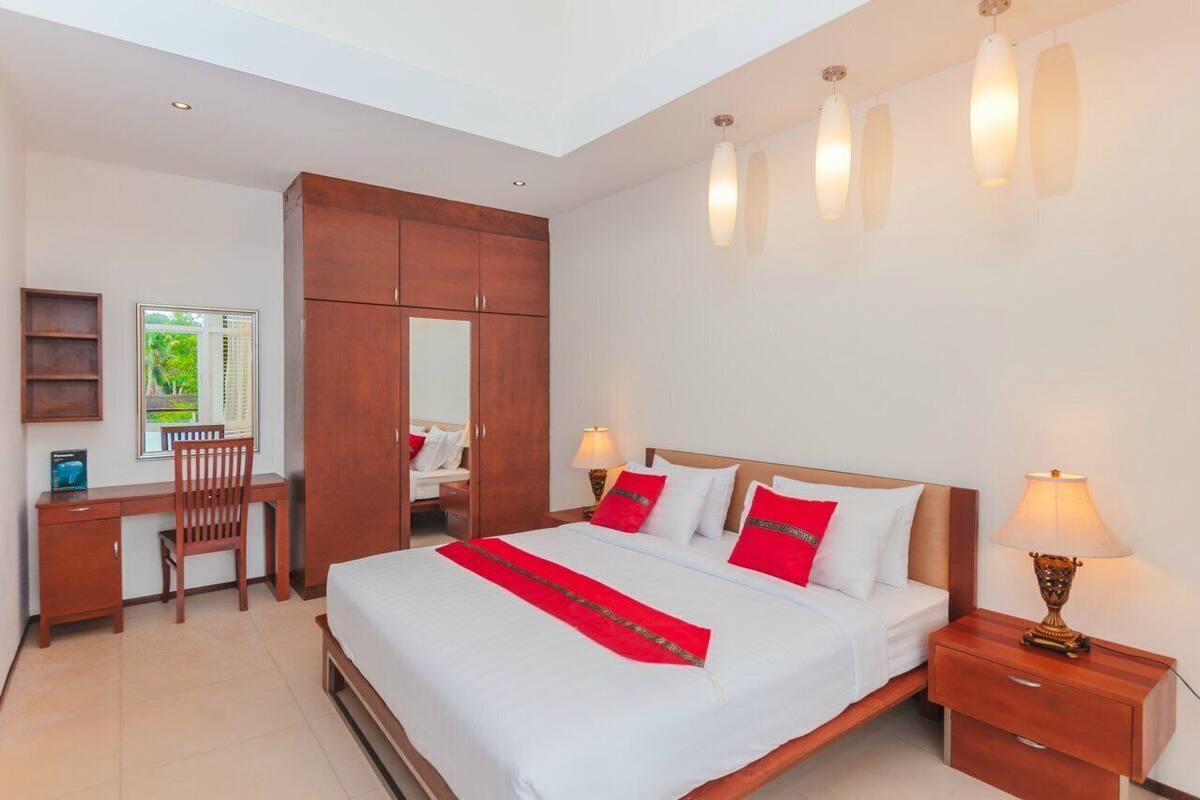 A hotel room with a large bed, a desk, a chair, a wardrobe, and a window with a view of greenery.