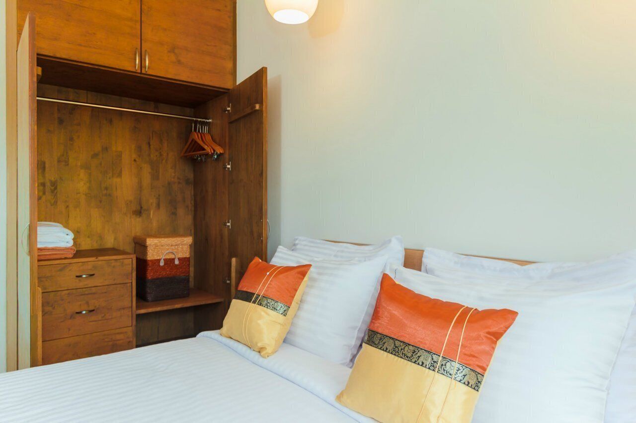 A bedroom with a wooden wardrobe, a bed with white bedding, and a bedside table with a basket and folded towels.