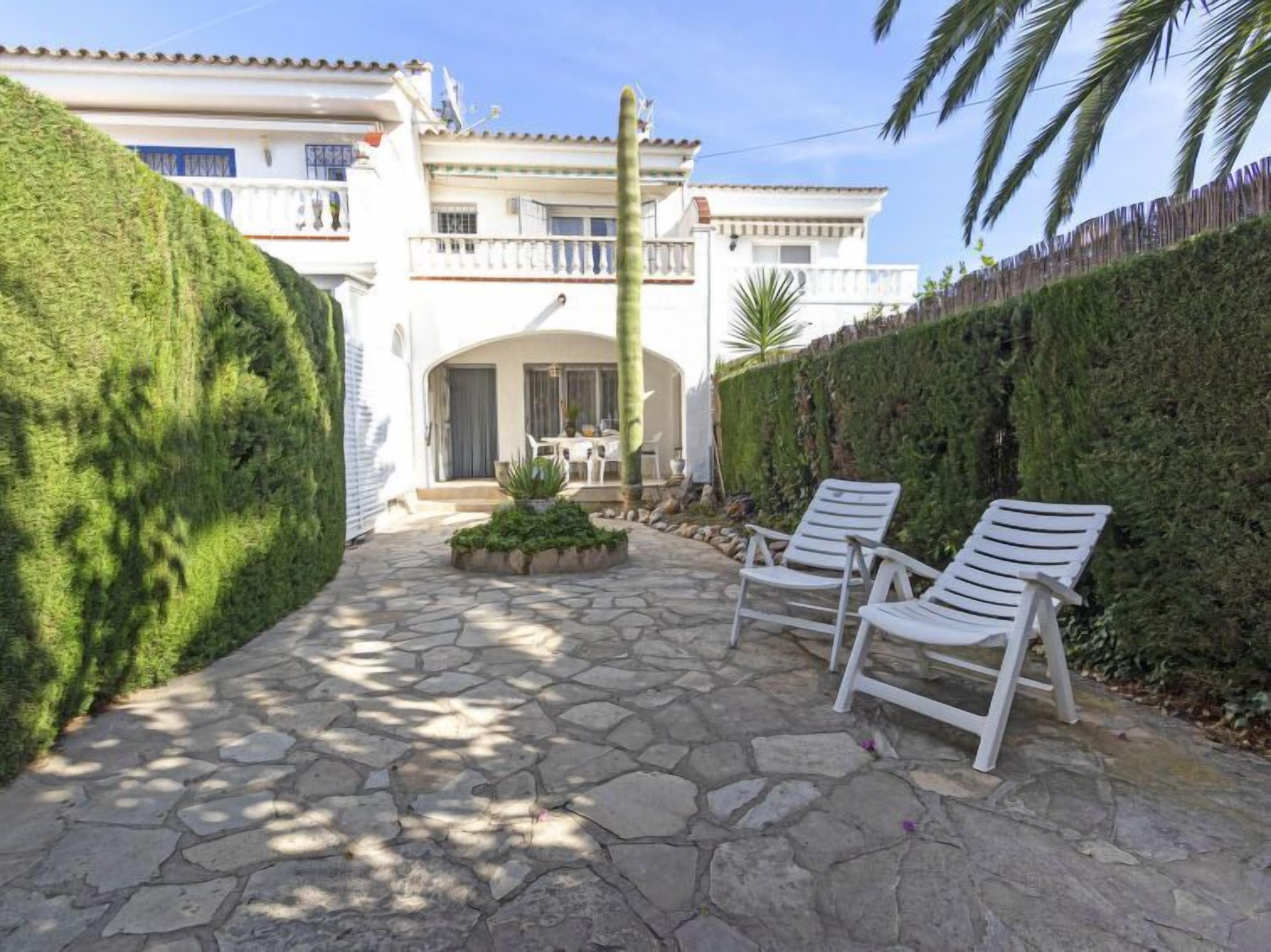 A stone-paved patio with white lounge chairs, a small garden, and a white building with balconies.