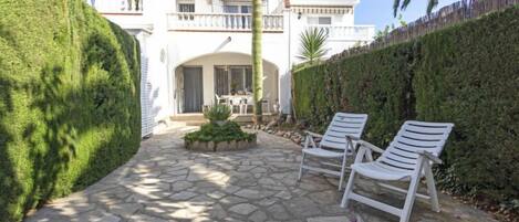 A stone-paved patio with white lounge chairs, a small garden, and a white building with balconies.