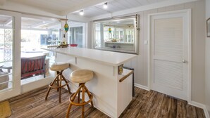 Une cuisine avec un plan de travail blanc, deux tabourets, un grand miroir et une porte à panneaux ajourés.