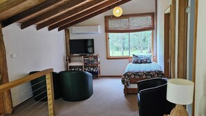 A cozy attic bedroom with a sloped ceiling, a bed with a floral duvet, a black armchair, a wooden side table with a lamp, and a television on a stand. - TRANQUIL BUSH RETREAT with
aircon wifi and indoor and outside fires (Brunkerville)