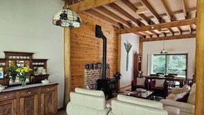 A cozy living room with a fireplace, white leather sofas, a wooden ceiling, and a dining area with natural light. - TRANQUIL BUSH RETREAT with
aircon wifi and indoor and outside fires (Brunkerville)