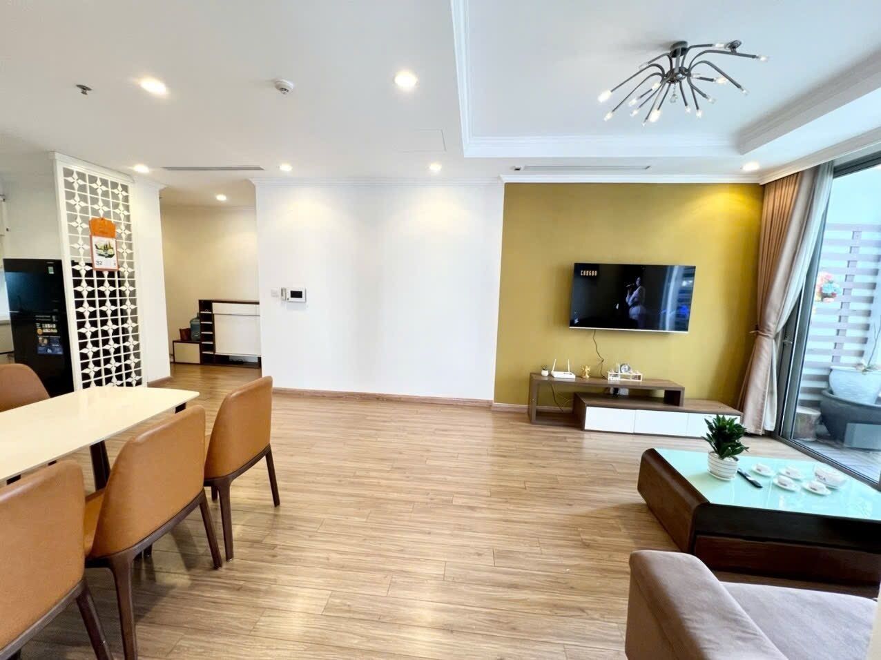 A modern living room with a dining area, a flat-screen TV mounted on the wall, and a large window with curtains.