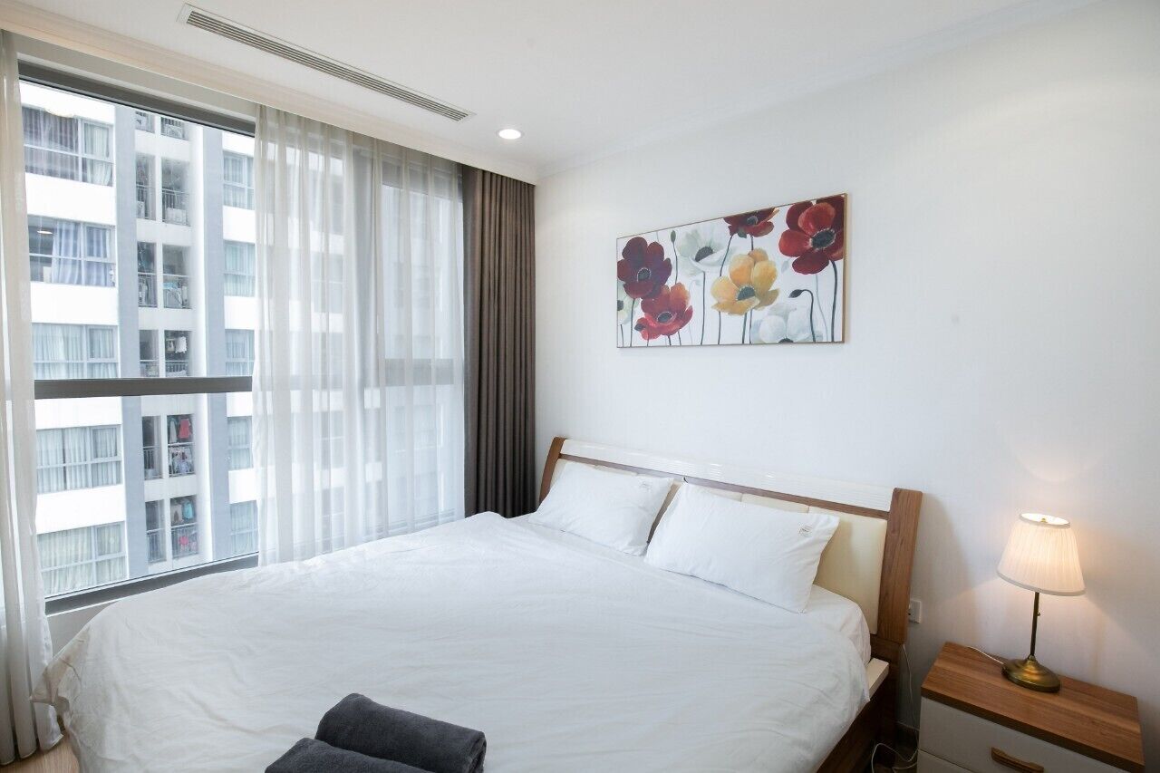 Une chambre d’hôtel avec un grand lit, une table de chevet avec une lampe, une fenêtre avec des rideaux et un tableau représentant des fleurs accroché au mur.