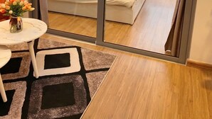 A modern bedroom with a large sliding glass door leading to a balcony, a bed with white linens, a small round table with a vase of flowers, and a patterned rug. - Bom Homestay Vinhomes Times City (Hanoi)