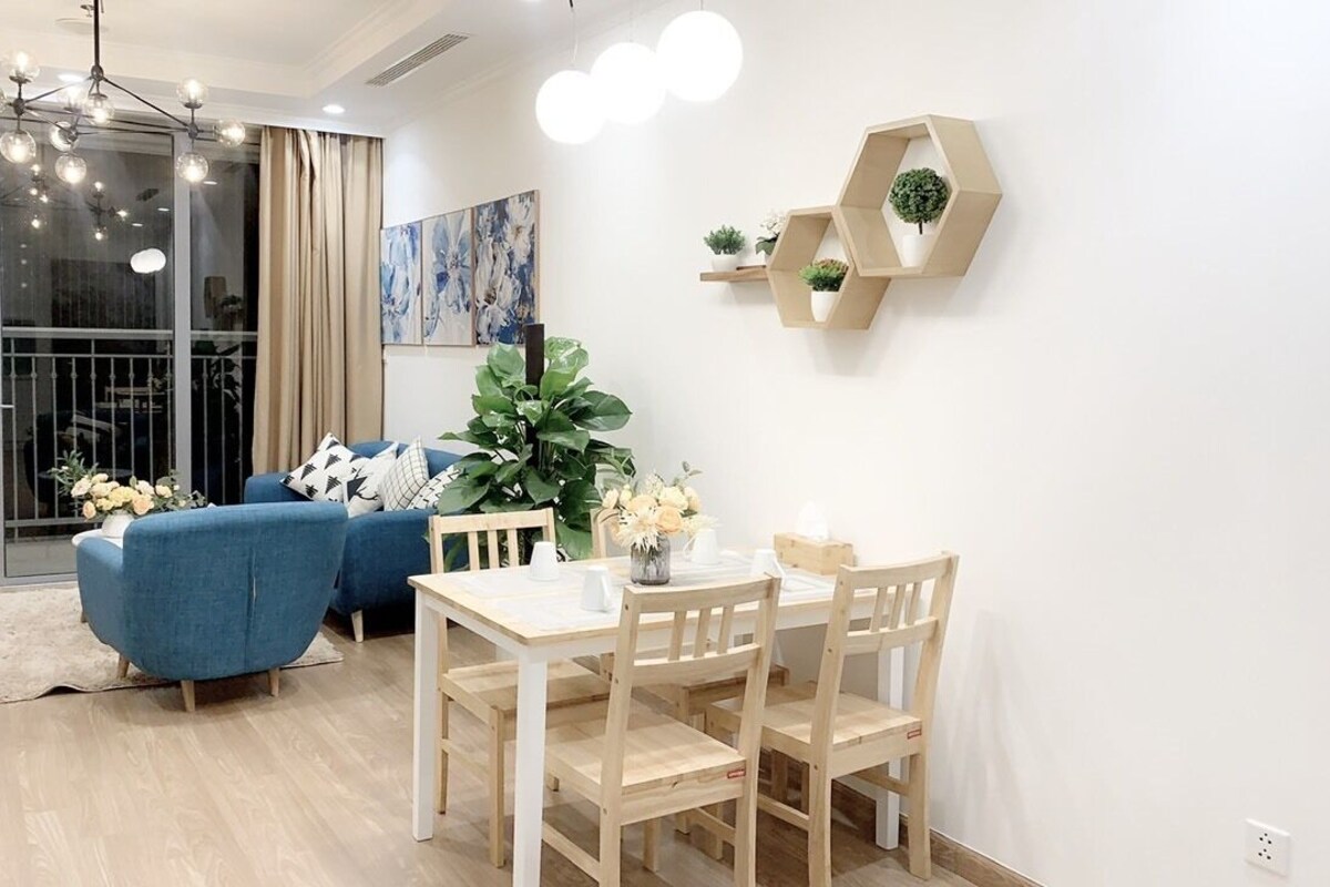 A modern living room with a blue sofa, wooden dining table, and chairs. There is a large plant and artwork on the wall.