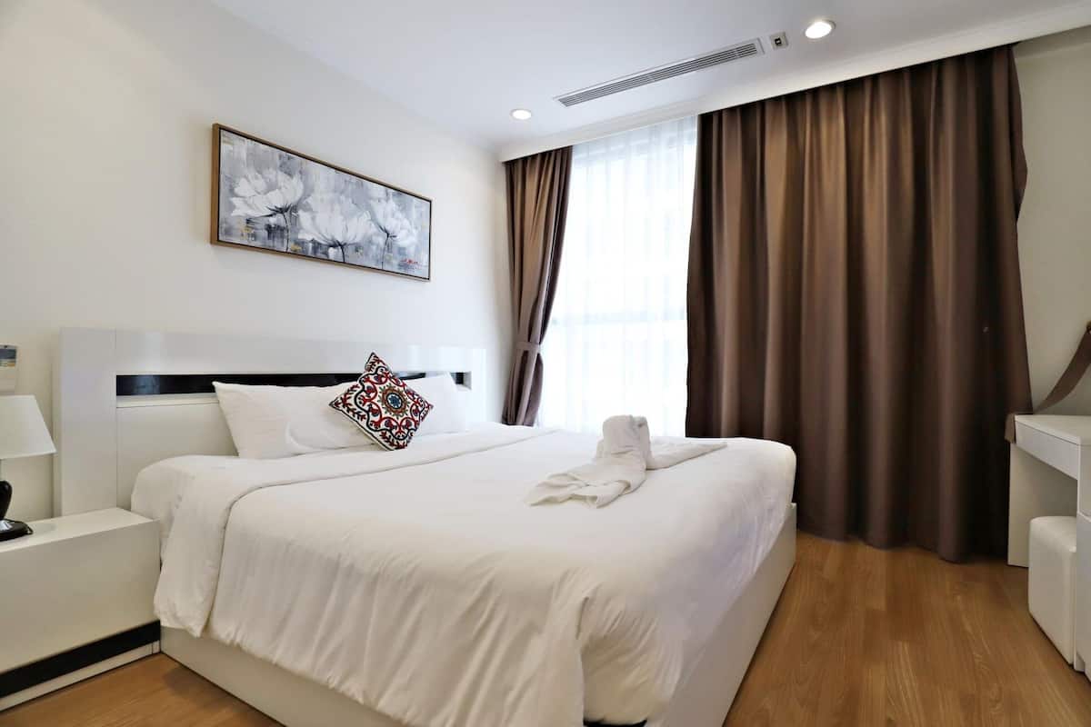 A hotel room with a large bed, a bedside table with a lamp, a wall-mounted artwork, and a window with curtains.
