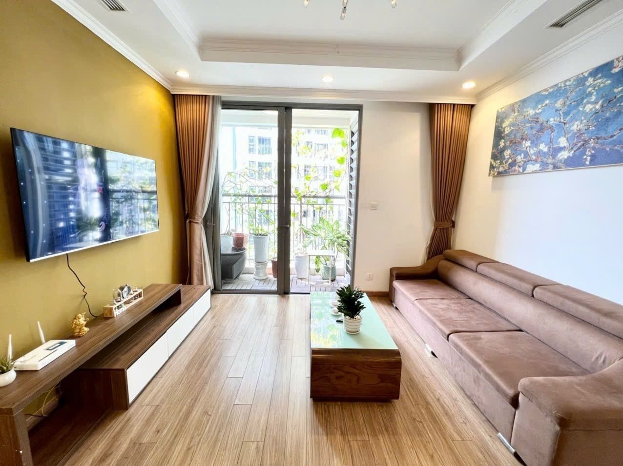A modern living room with a flat-screen TV, a sofa, a coffee table, and a sliding glass door leading to a balcony with plants.