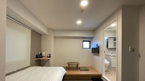 Habitación de hotel con cama, una mesita con cojín verde, televisor montado en la pared y baño visible a través de una puerta abierta.