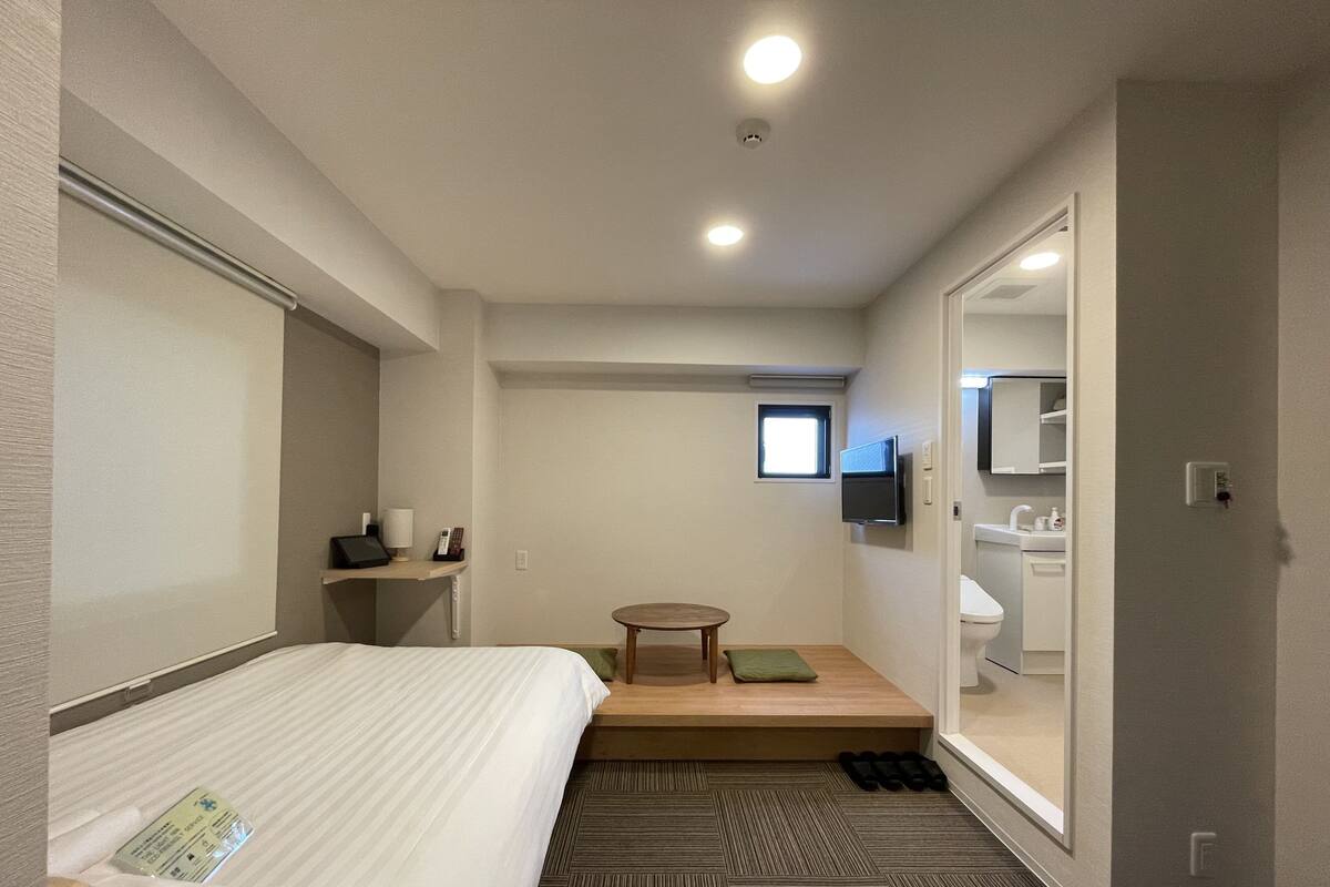 Ein Hotelzimmer mit einem Bett, einem kleinen Tisch mit einem grünen Kissen, einem an der Wand montierten Fernseher und einem durch eine offene Tür einsehbaren Badezimmer.