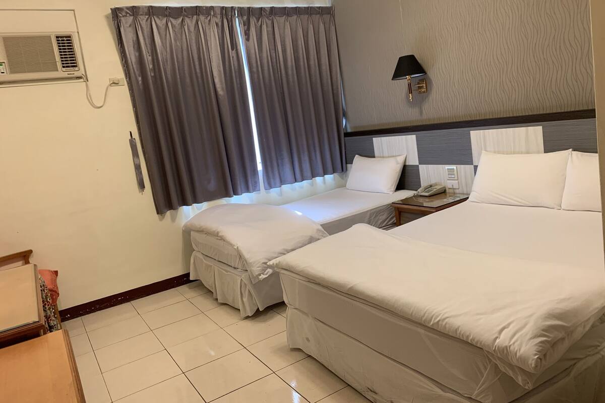 A hotel room with two beds, a bedside table with a lamp, a telephone, and a window with curtains.