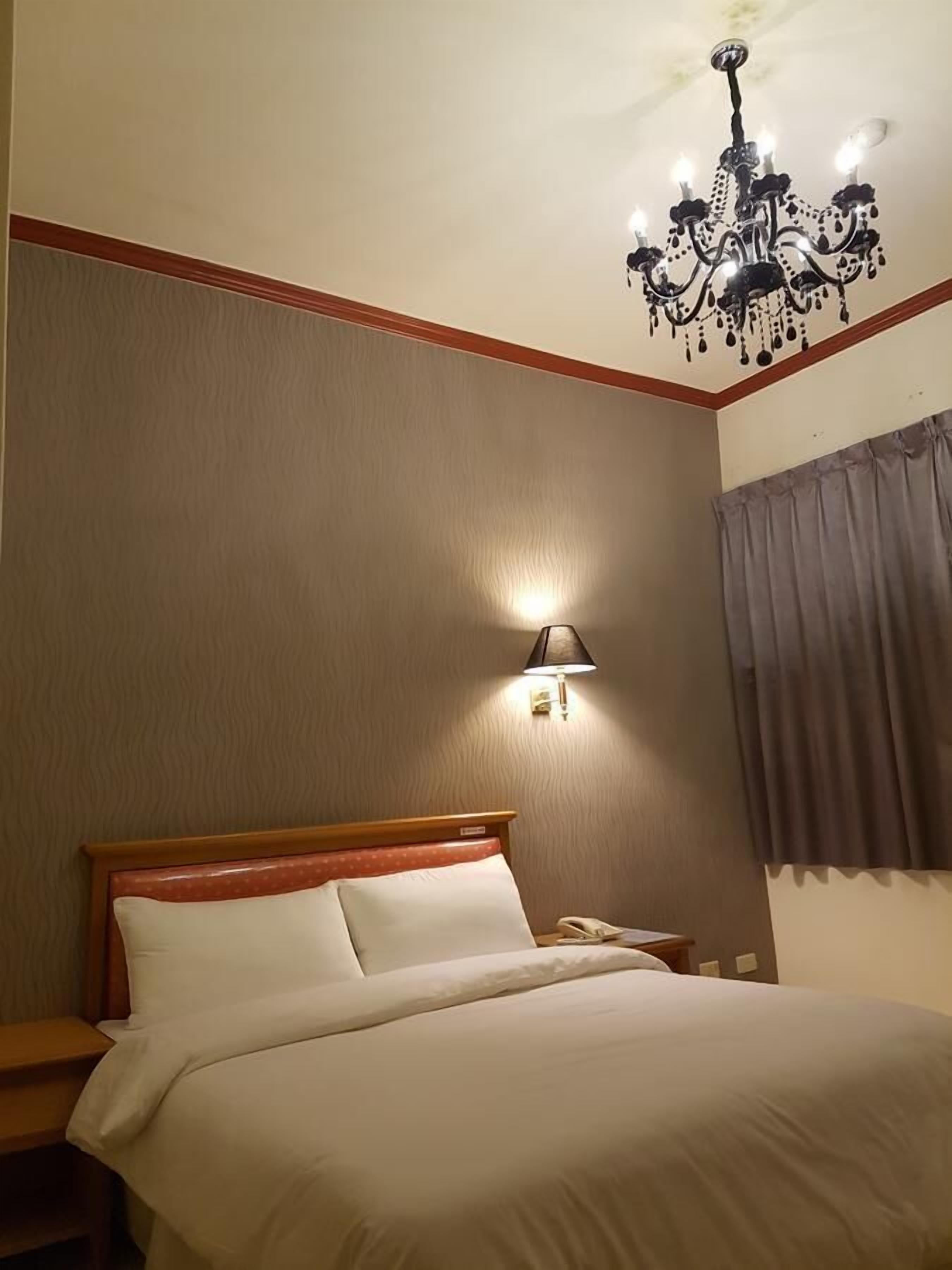 A hotel room with a bed, a nightstand, a telephone, a wall-mounted lamp, and a chandelier.