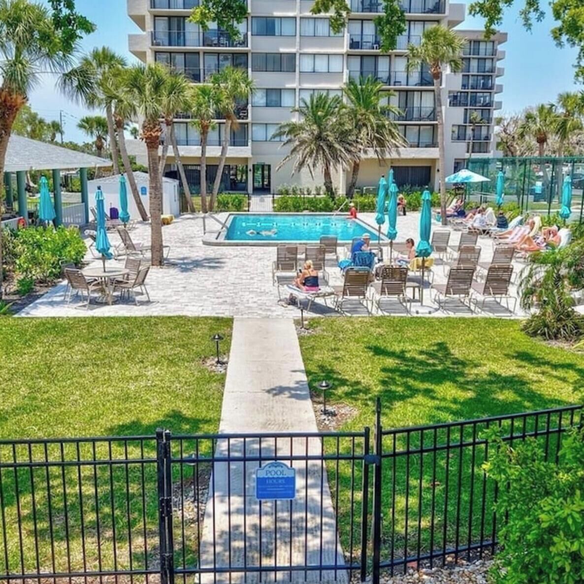 A hotel pool area with lounge chairs and umbrellas, surrounded by palm trees and a building with balconies.