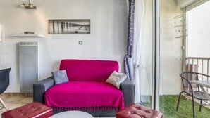 Una habitación con un sofá rojo brillante, dos otomanas rojas, una mesa redonda blanca y un sillón gris. Hay un cuadro en la pared, un calentador y un balcón con una silla.