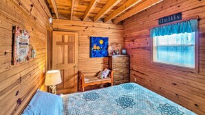 Ett mysigt rum med träväggar, en säng med ett blått täcke, en byrå, ett nattduksbord, en lampa och en tavla på väggen.