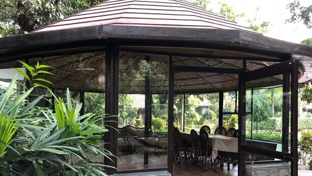 Un espace extérieur abrité, aménagé pour les repas, avec des murs en pierre et des panneaux de verre, le tout entouré de verdure.