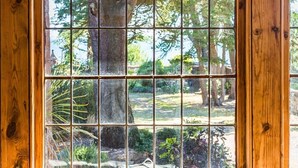 Una ventana de madera con diseño de cuadrícula, un banco con cojines estampados y vistas a árboles y follaje.