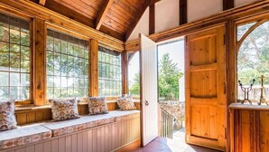 Interior de madera con un banco junto a la ventana, una puerta que da al exterior y una pared de piedra visible a través de la ventana.
