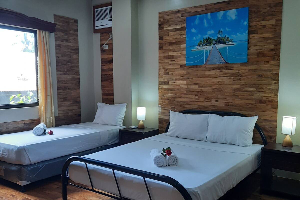 A hotel room with two beds, wooden flooring, a painting on the wall, and a window with curtains.