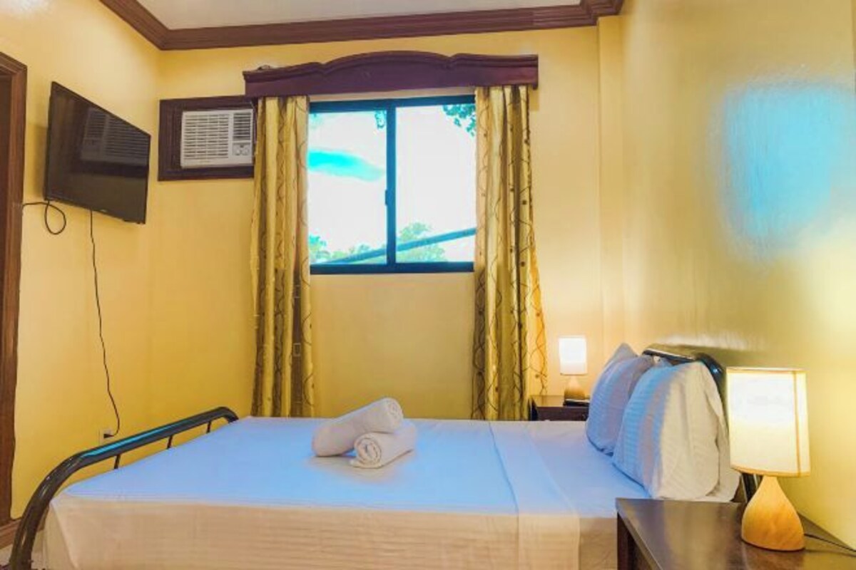 Habitación de hotel con cama, televisor en la pared, ventana con cortinas y lámpara de noche.