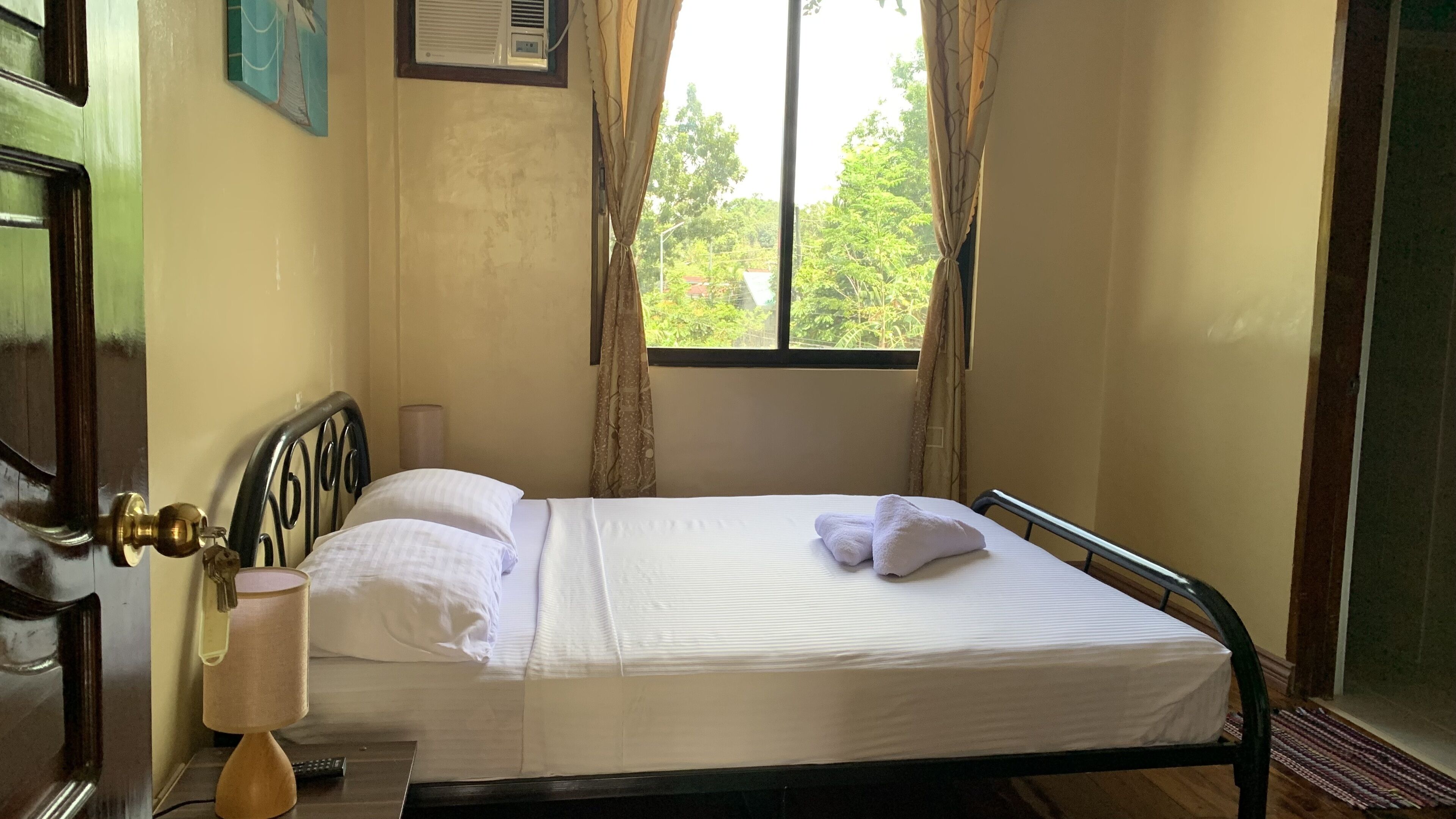 A single bed with white linens and a pillow, a bedside lamp, and a window with curtains.