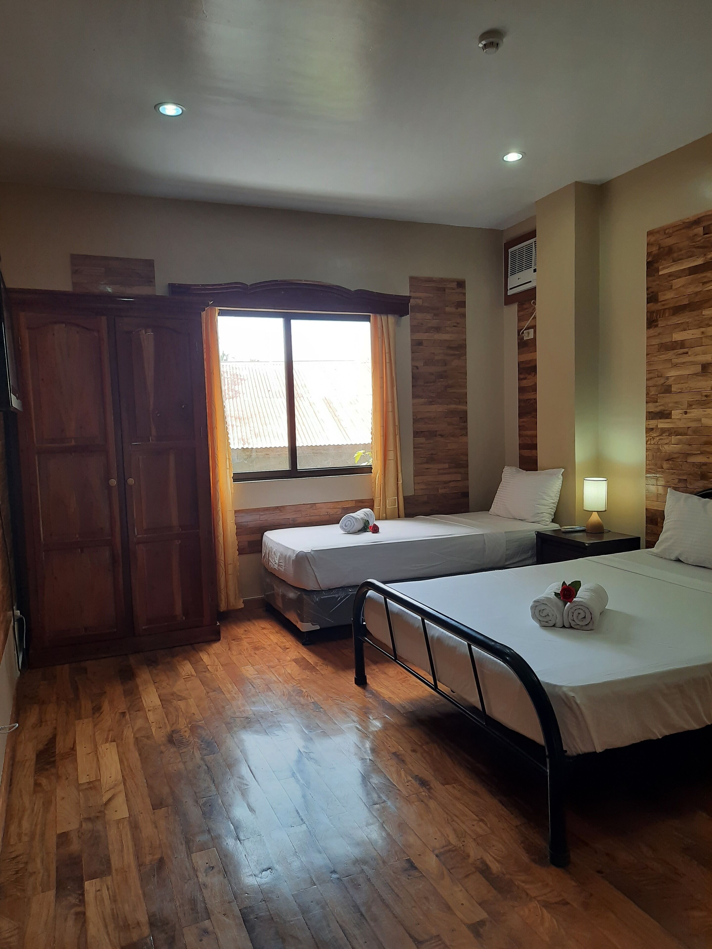 Camera d'albergo con due letti, pavimento in legno, un'ampia finestra con tende e pannello decorativo in legno.