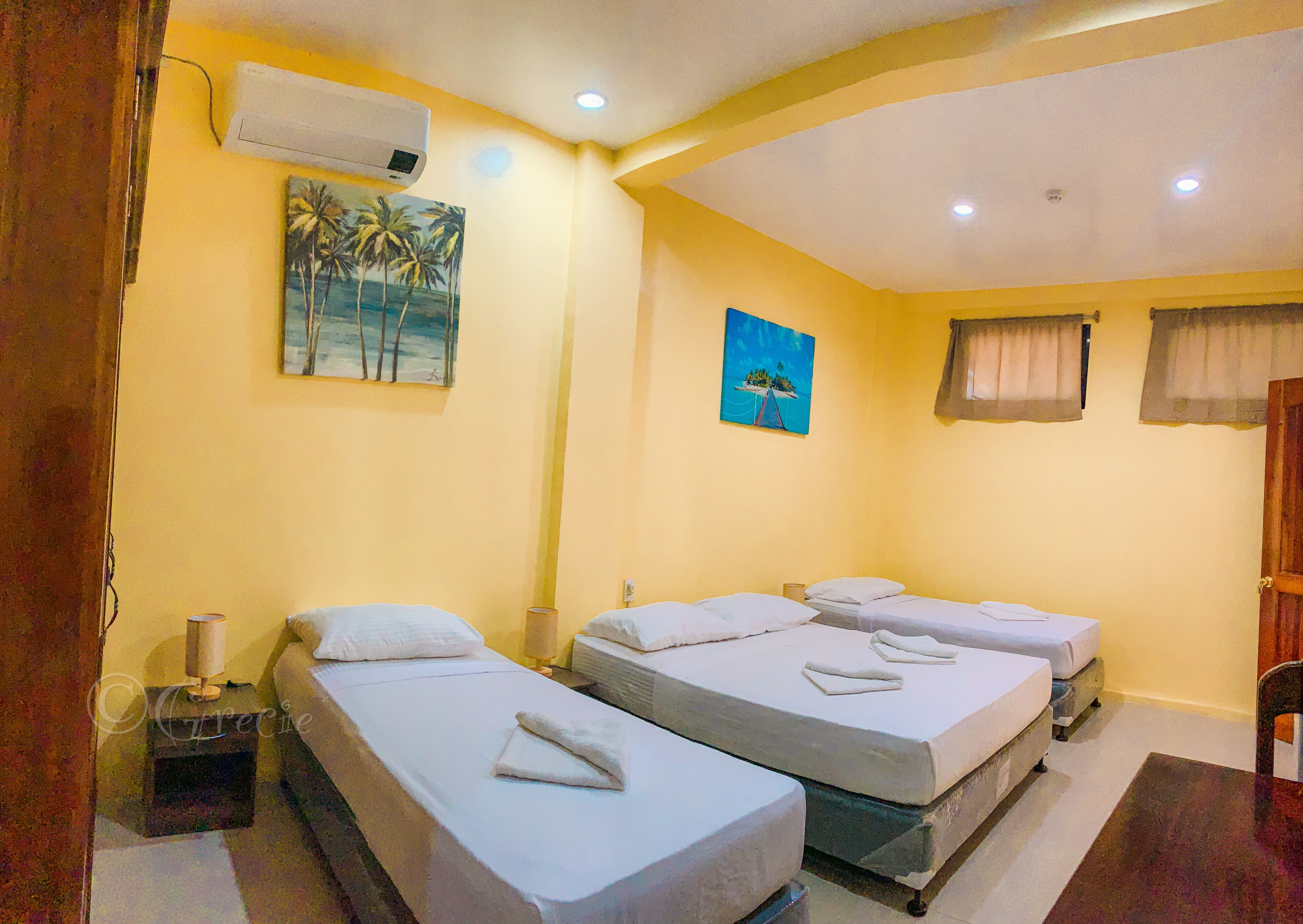Quarto de hotel com três camas de solteiro, uma pintura de palmeiras e um aparelho de ar condicionado na parede.