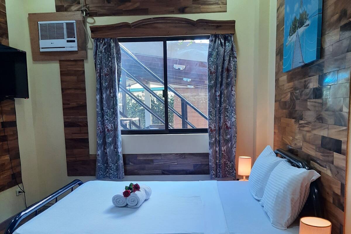 A hotel room with a bed, a window with a view of a building, a mounted air conditioner, and a painting on the wall.