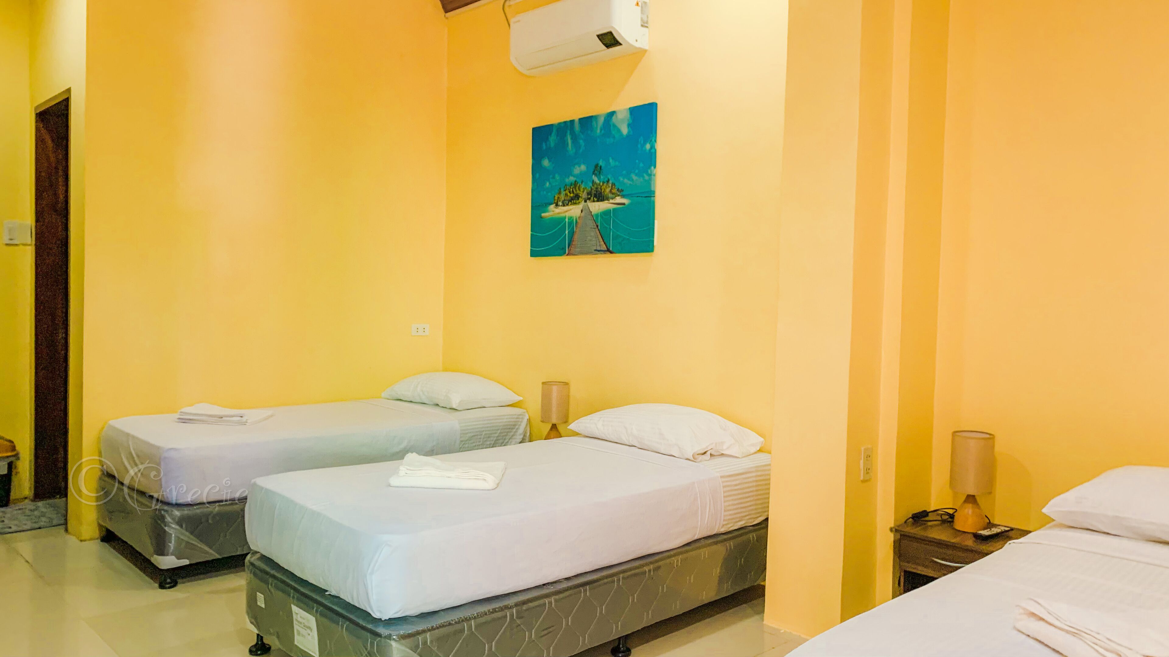 Habitación de hotel con dos camas individuales, un cuadro de una isla tropical en la pared y un equipo de aire acondicionado sobre las camas.